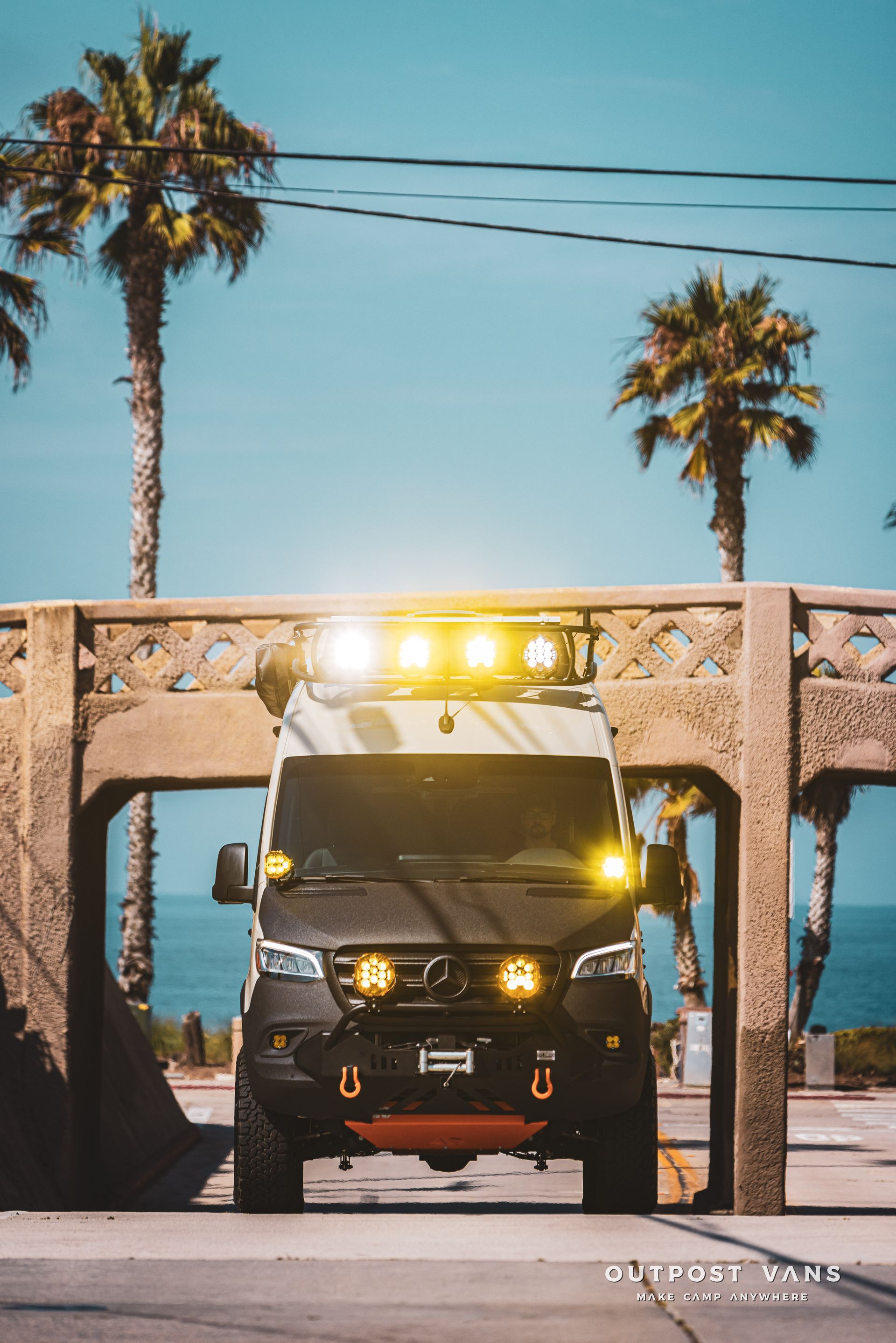 A van is parked under a bridge with palm trees in the background.