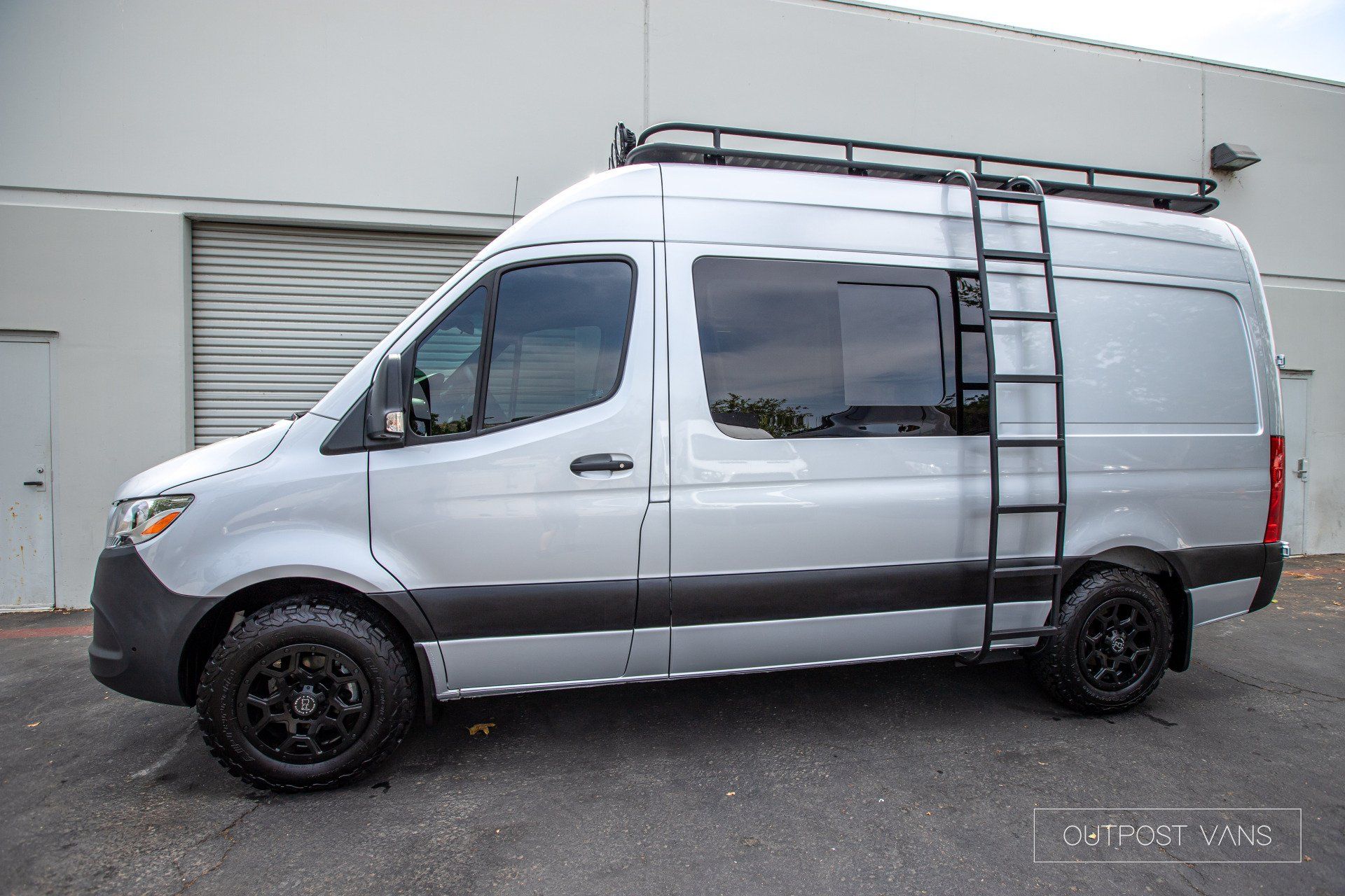 A silver van with a ladder on the side is parked in front of a building.