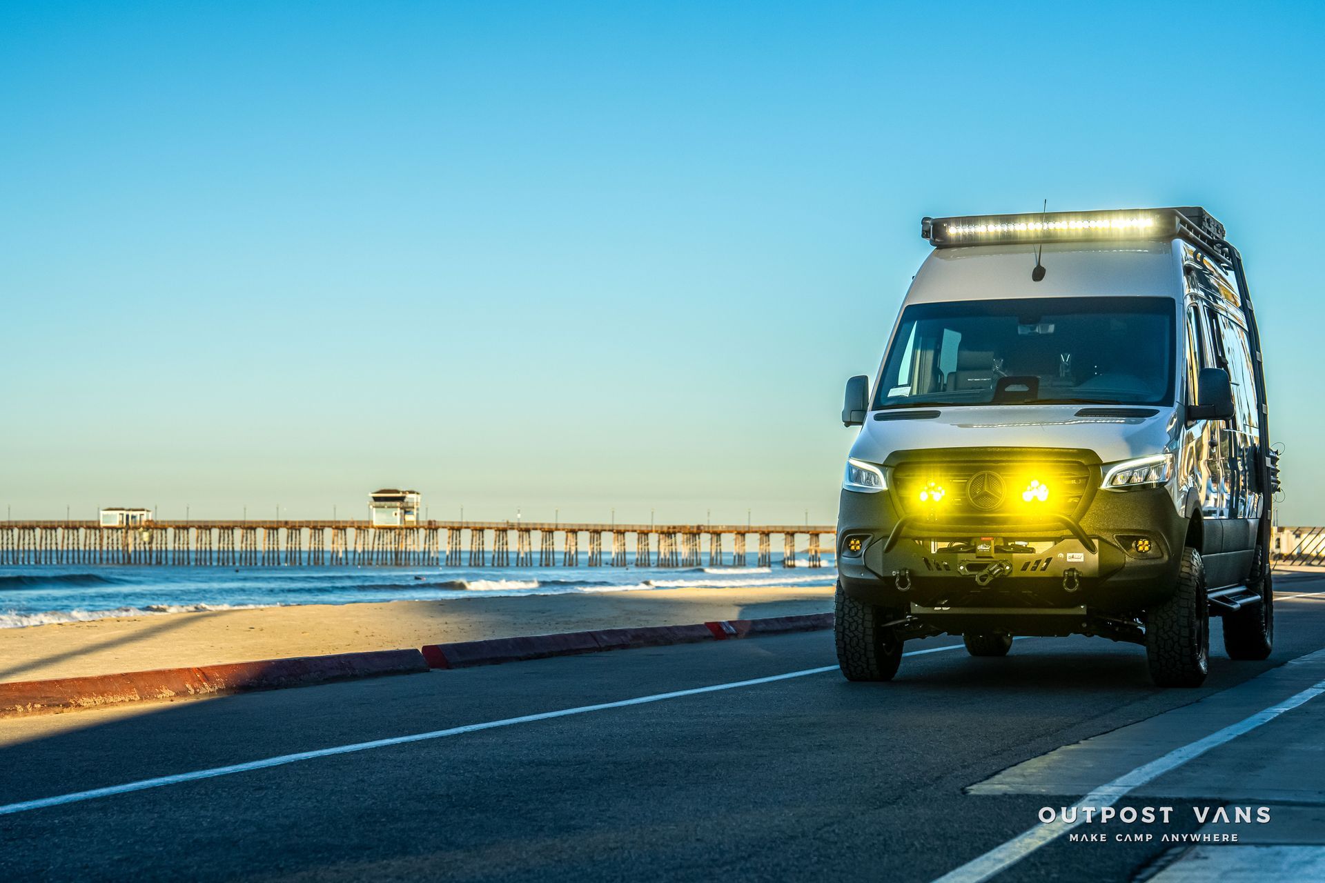 Silver van with yellow lights driving on a road next to the ocean, pier in background.
