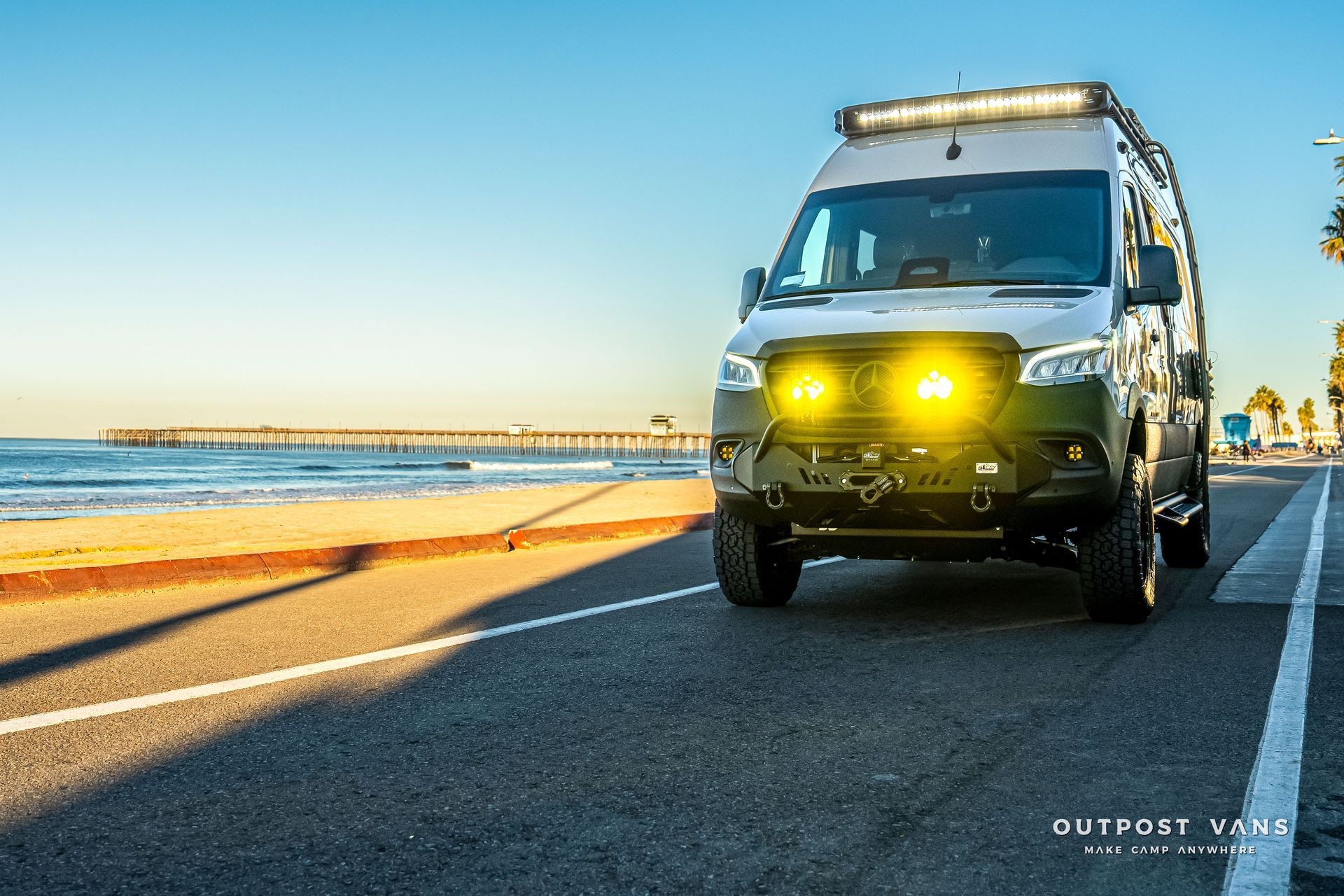 Off-road van with bright yellow lights parked on a road next to the beach, pier visible in the distance.