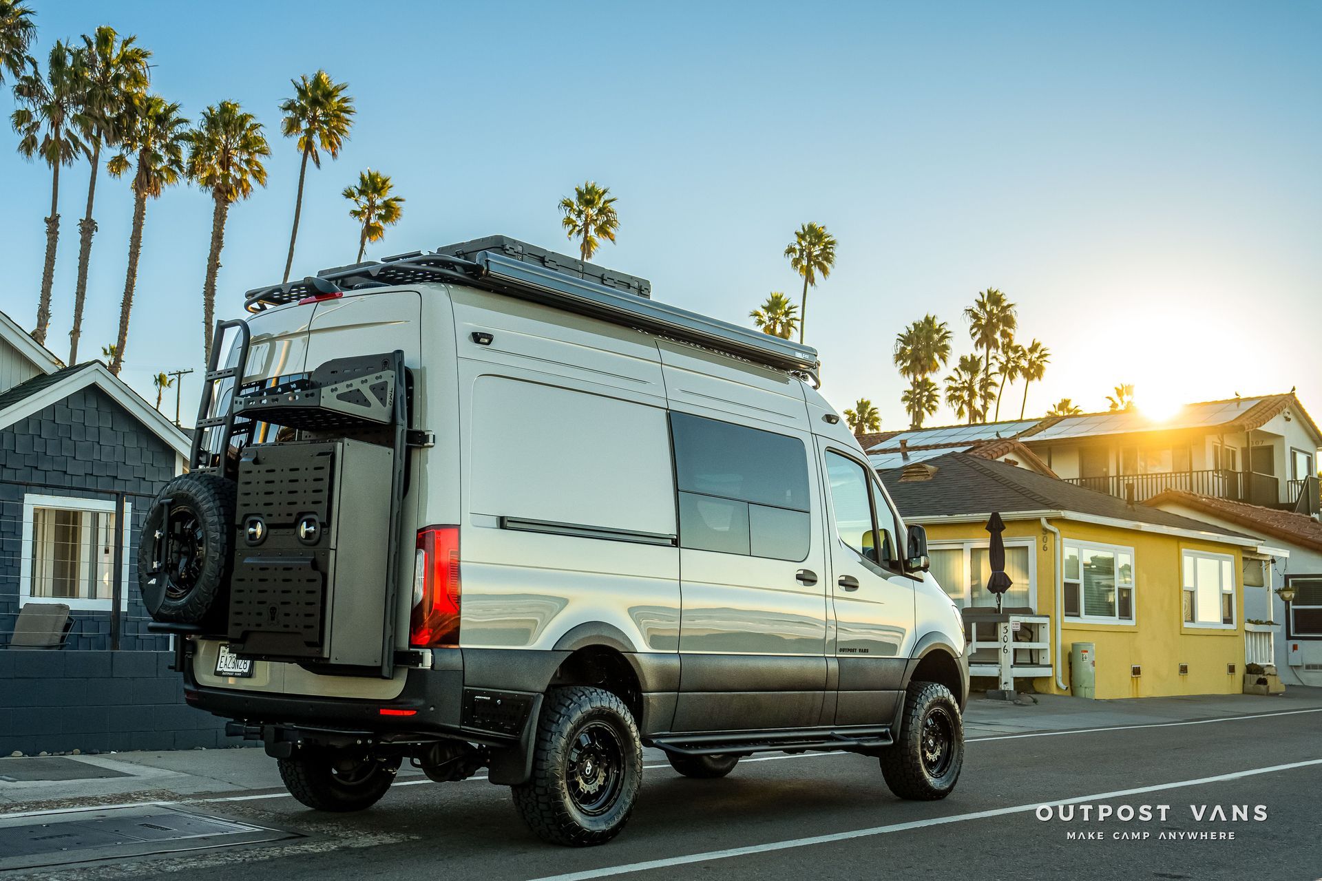 Gray campervan parked on a road by a beach. Palm trees and houses in the background.