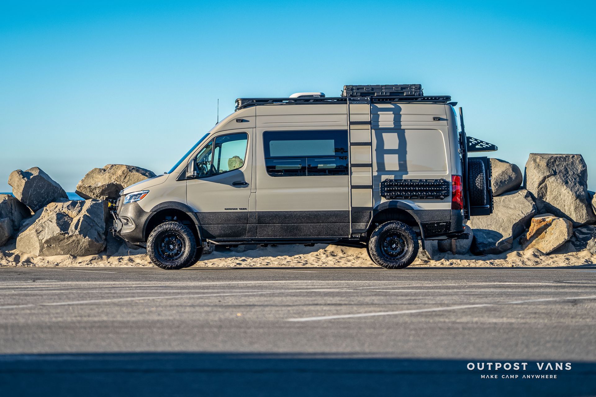 Gray and black camper van parked next to large rocks on a paved road near the ocean.