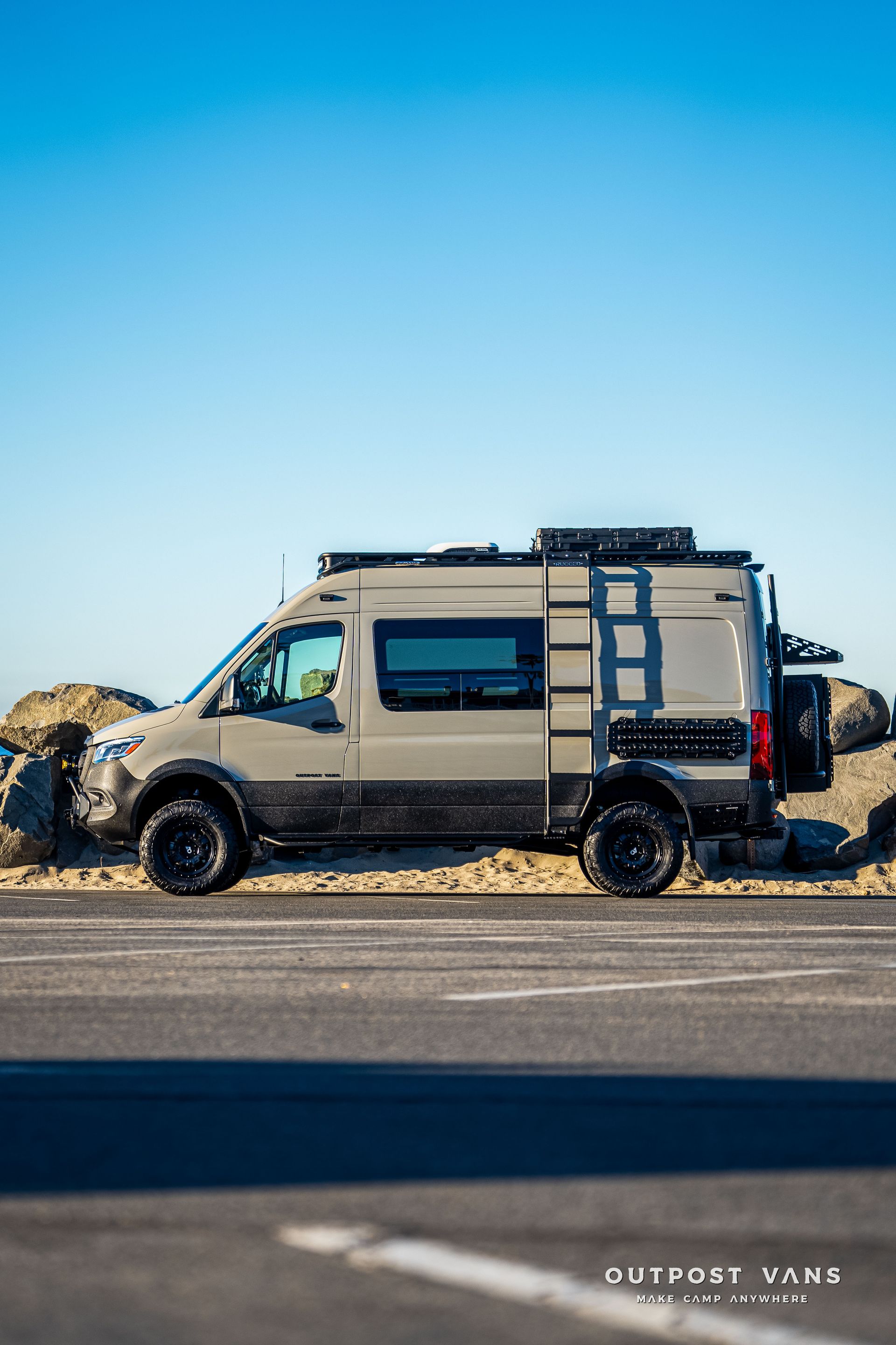 Gray camper van parked on asphalt, near rocks and ocean, under a blue sky.