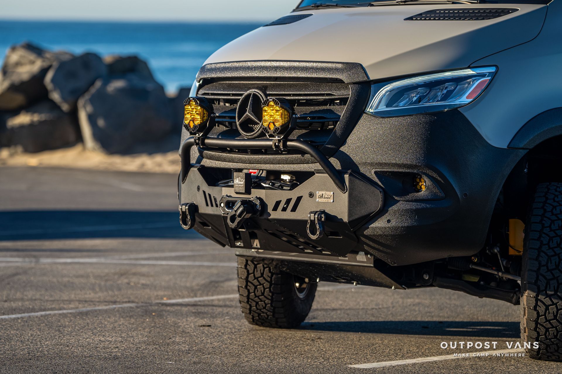 Modified Mercedes Sprinter van, olive-colored, with off-road bumper, lights, and winch, parked near the ocean.