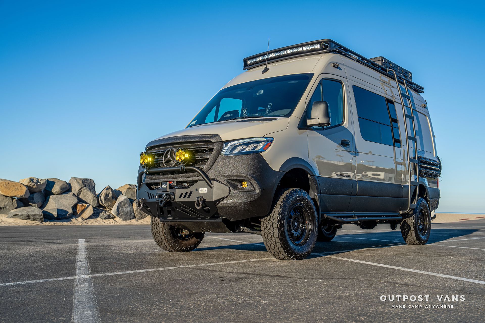 Tan and black off-road van on asphalt, equipped with lights and a roof rack against a blue sky.