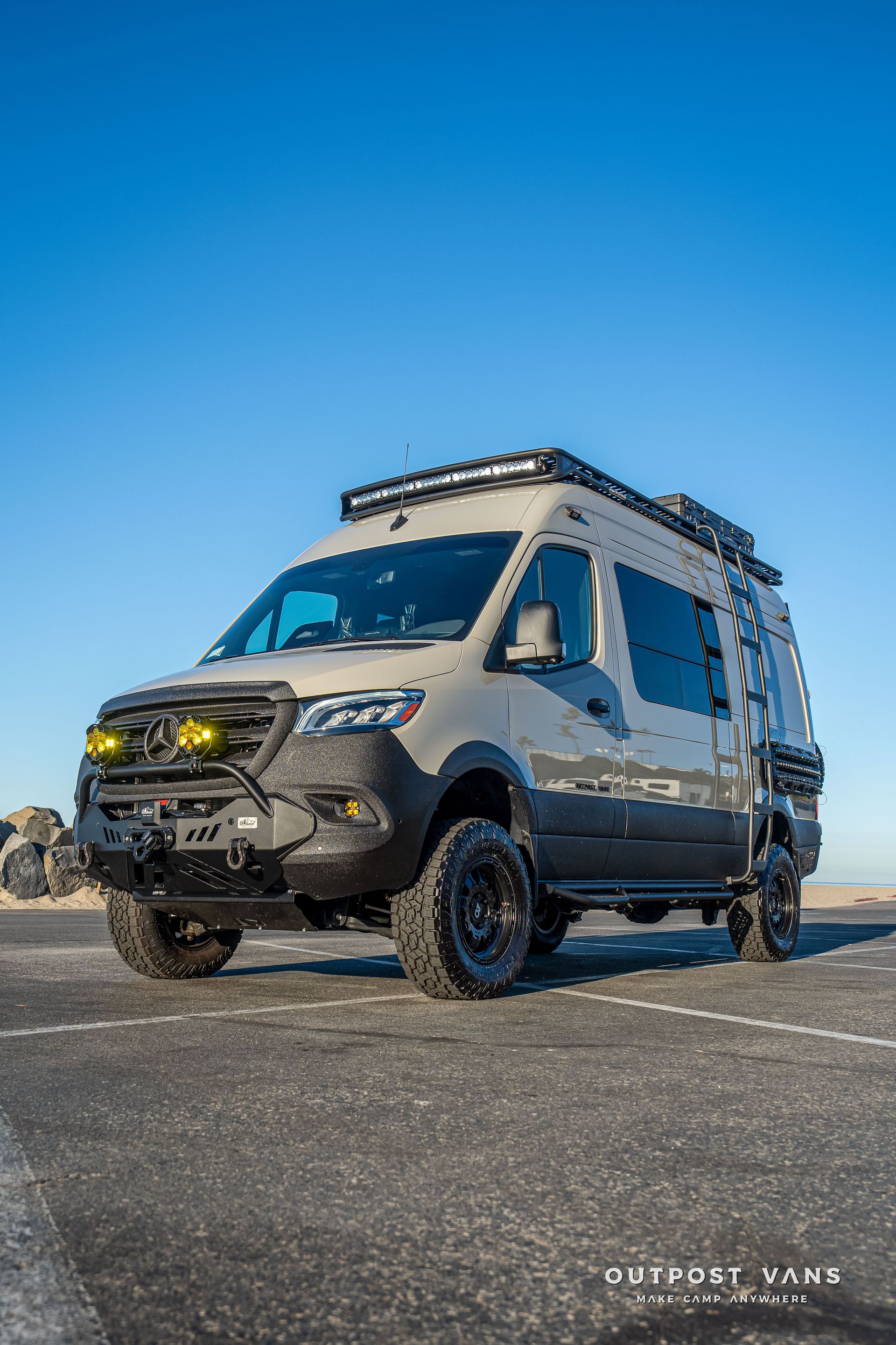 Grey off-road van with black accents, roof rack, and yellow fog lights parked on asphalt.