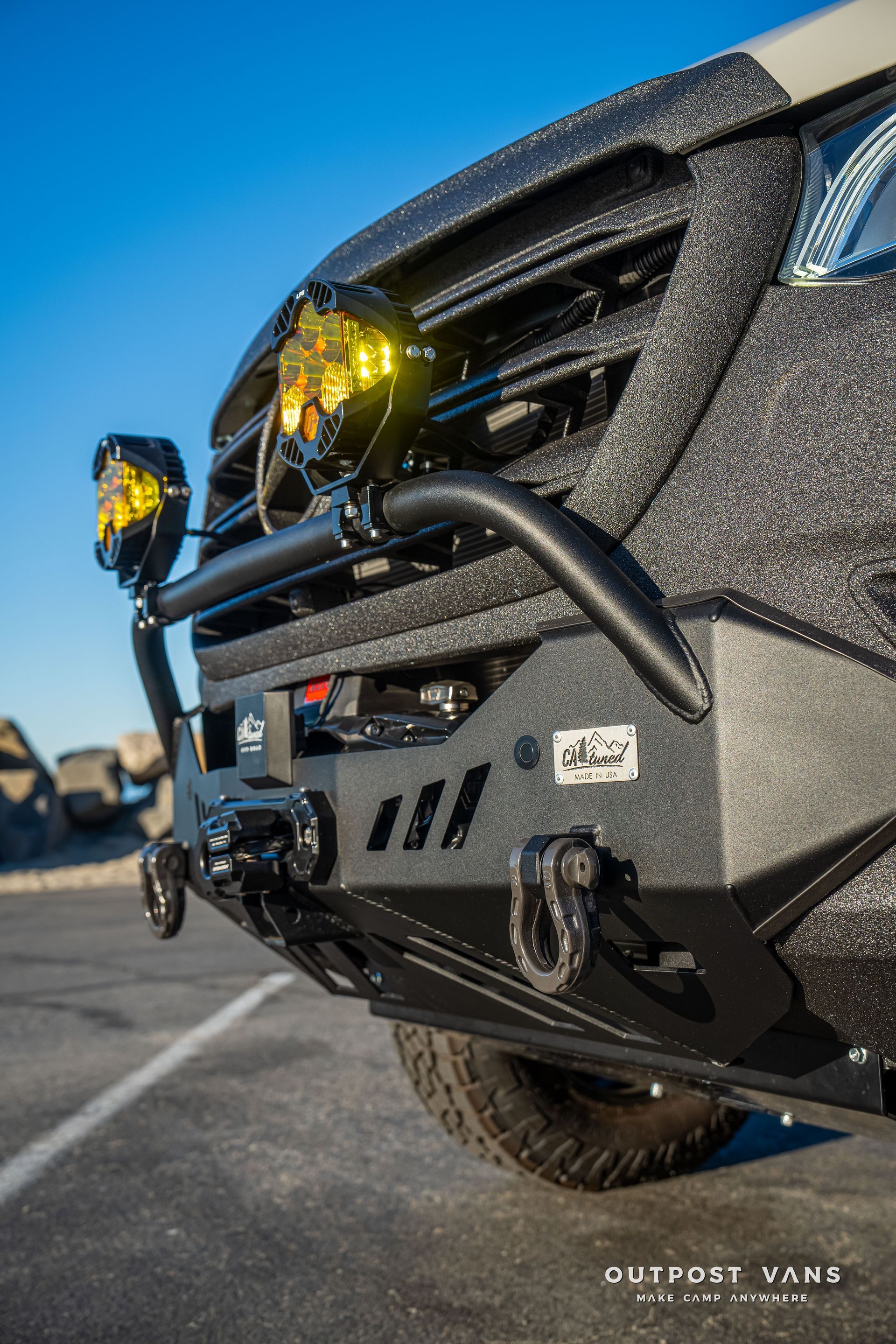 Black off-road vehicle front, rugged bumper, yellow spotlights, against a blue sky, text “Outpost Vans”.