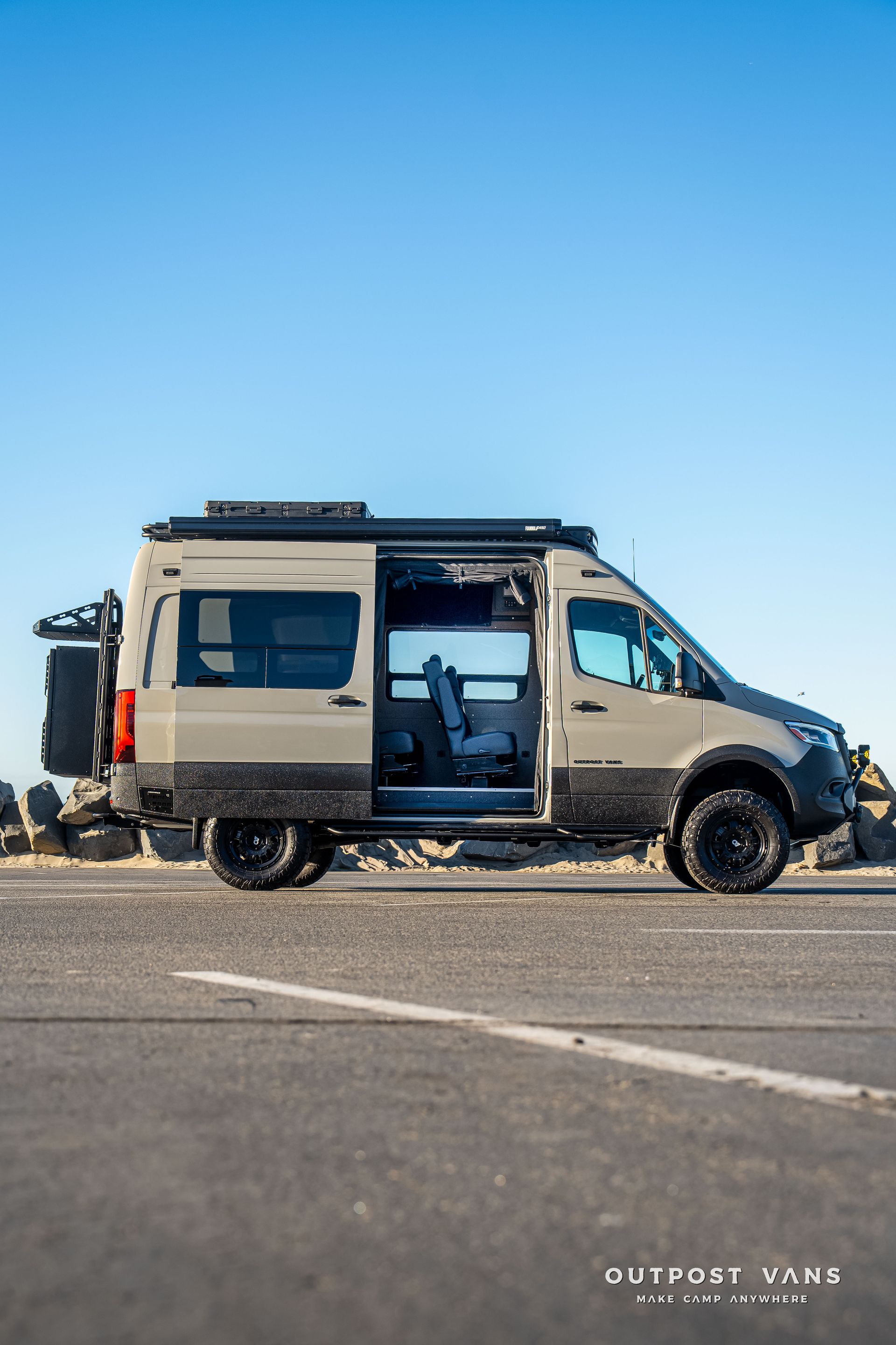 Tan and black campervan parked on a paved road with open sliding door. Blue sky.