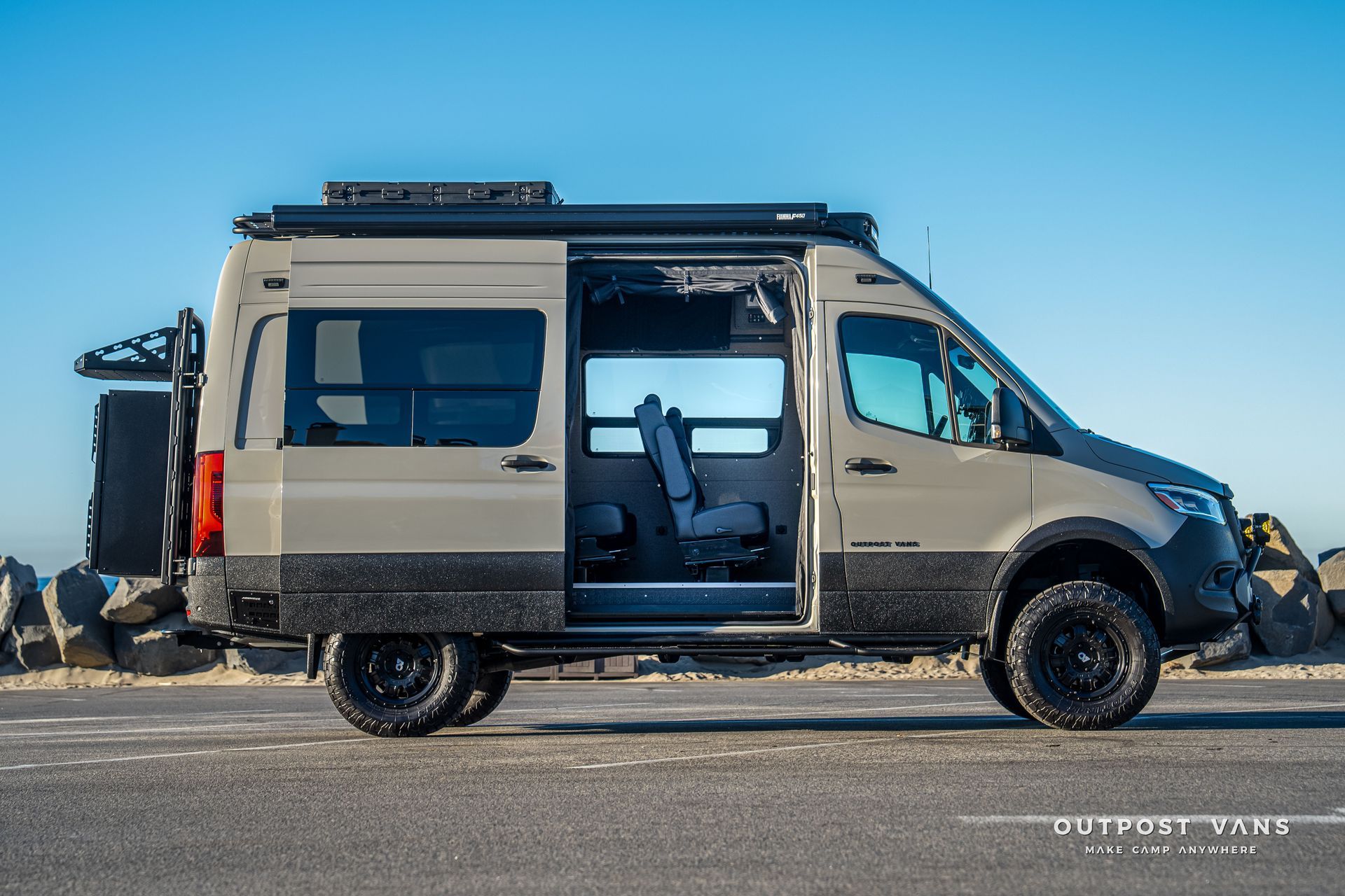 Off-road camper van with open sliding door. Tan and black, parked on a paved road next to rocks.