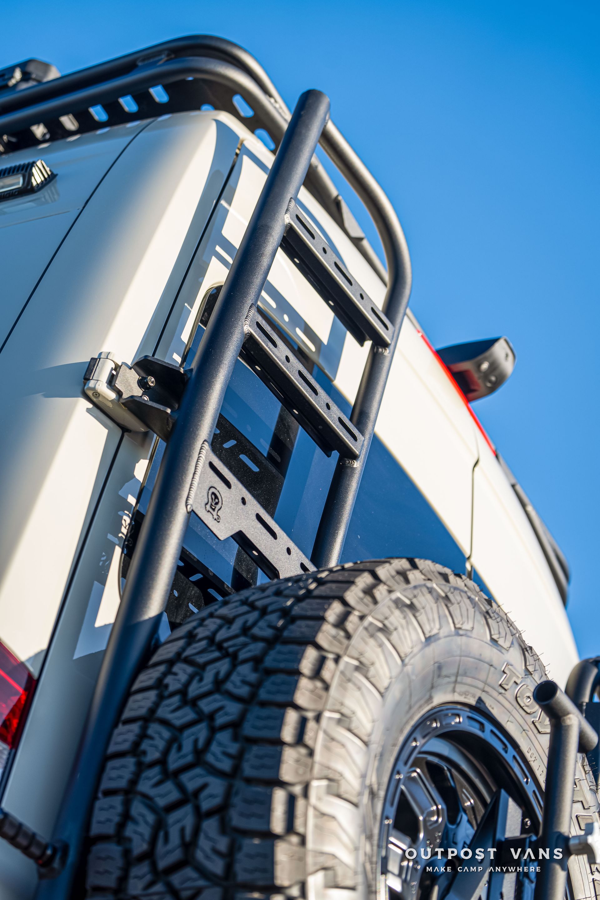Off-road vehicle with a black ladder, spare tire, and roof rack against a blue sky.
