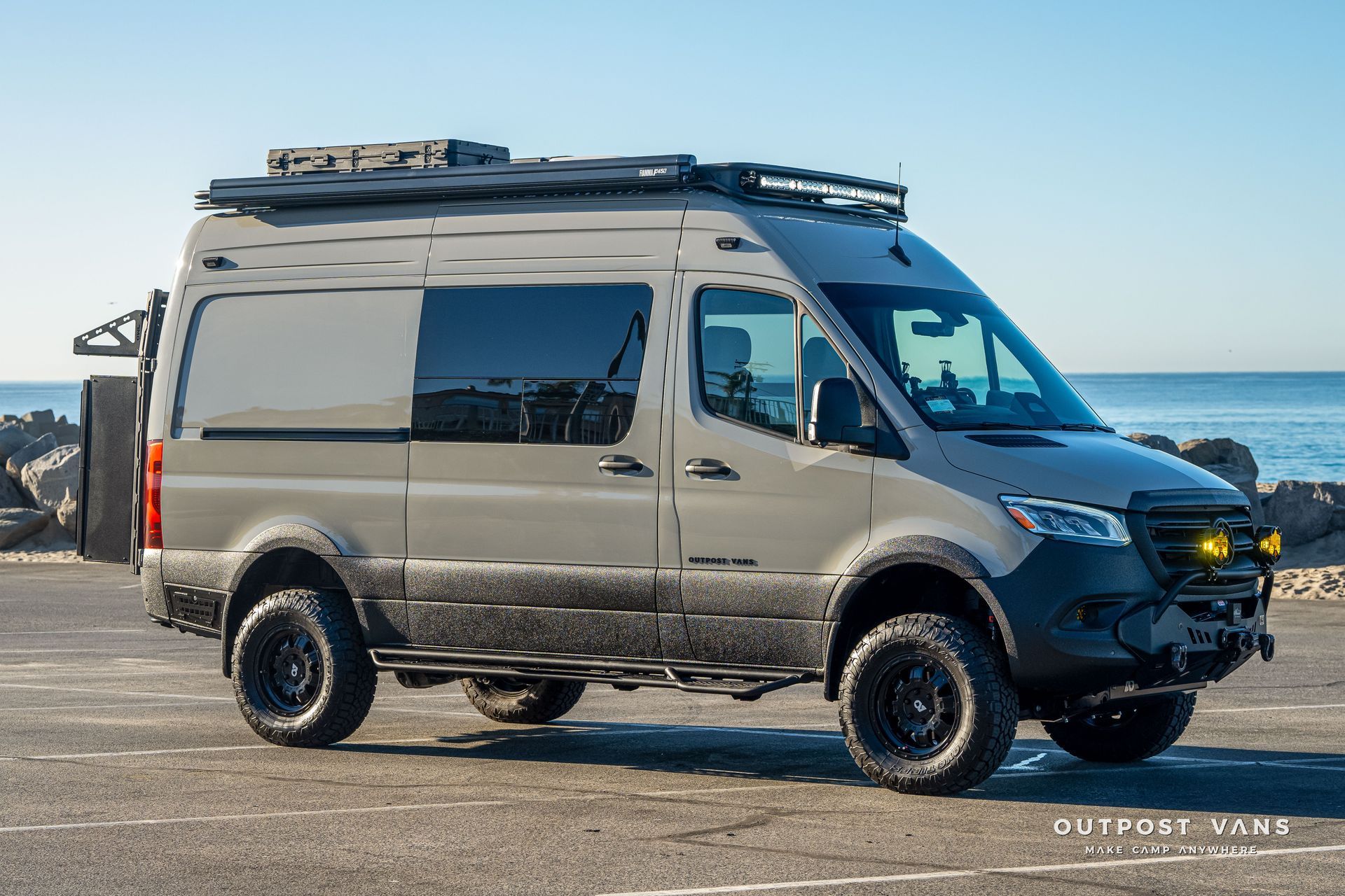 Gray camper van with black wheels, roof rack, and front bumper, parked near the ocean.