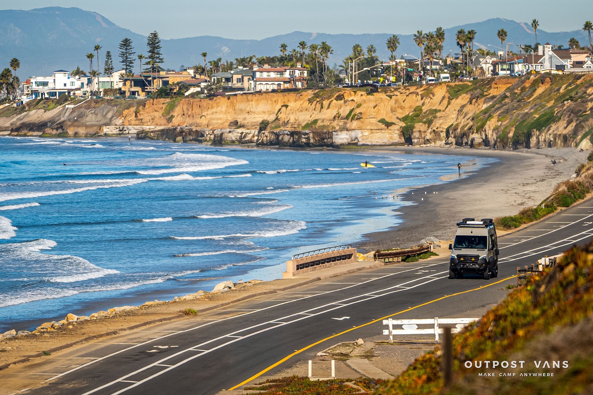 A van drives along a coastal road next to the ocean and beach, with houses on a cliff in the background.