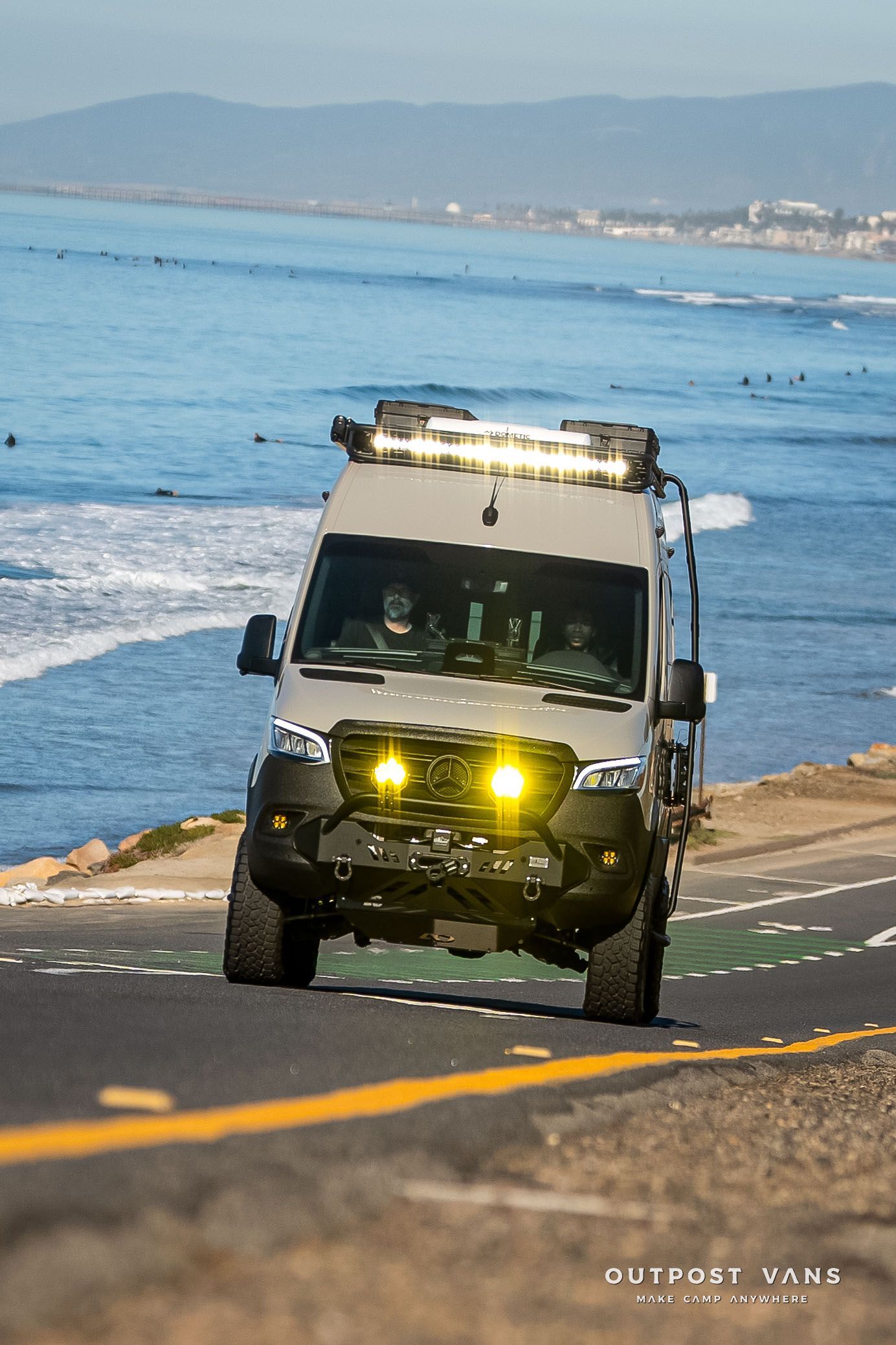 Off-road camper van driving on coastal road with ocean in the background, daytime.