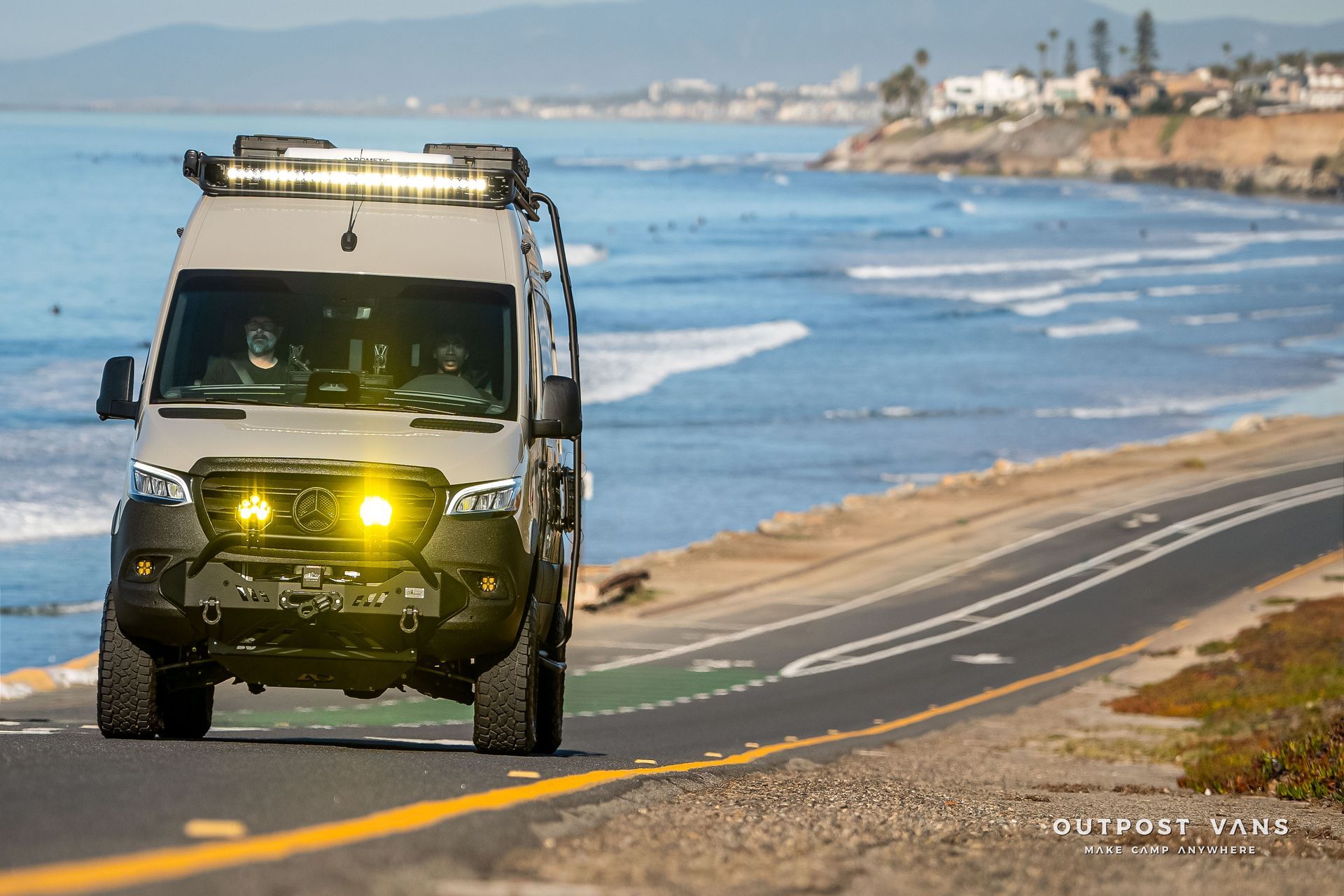 Off-road van drives on coastal road, bright yellow lights illuminate front. Ocean in background.
