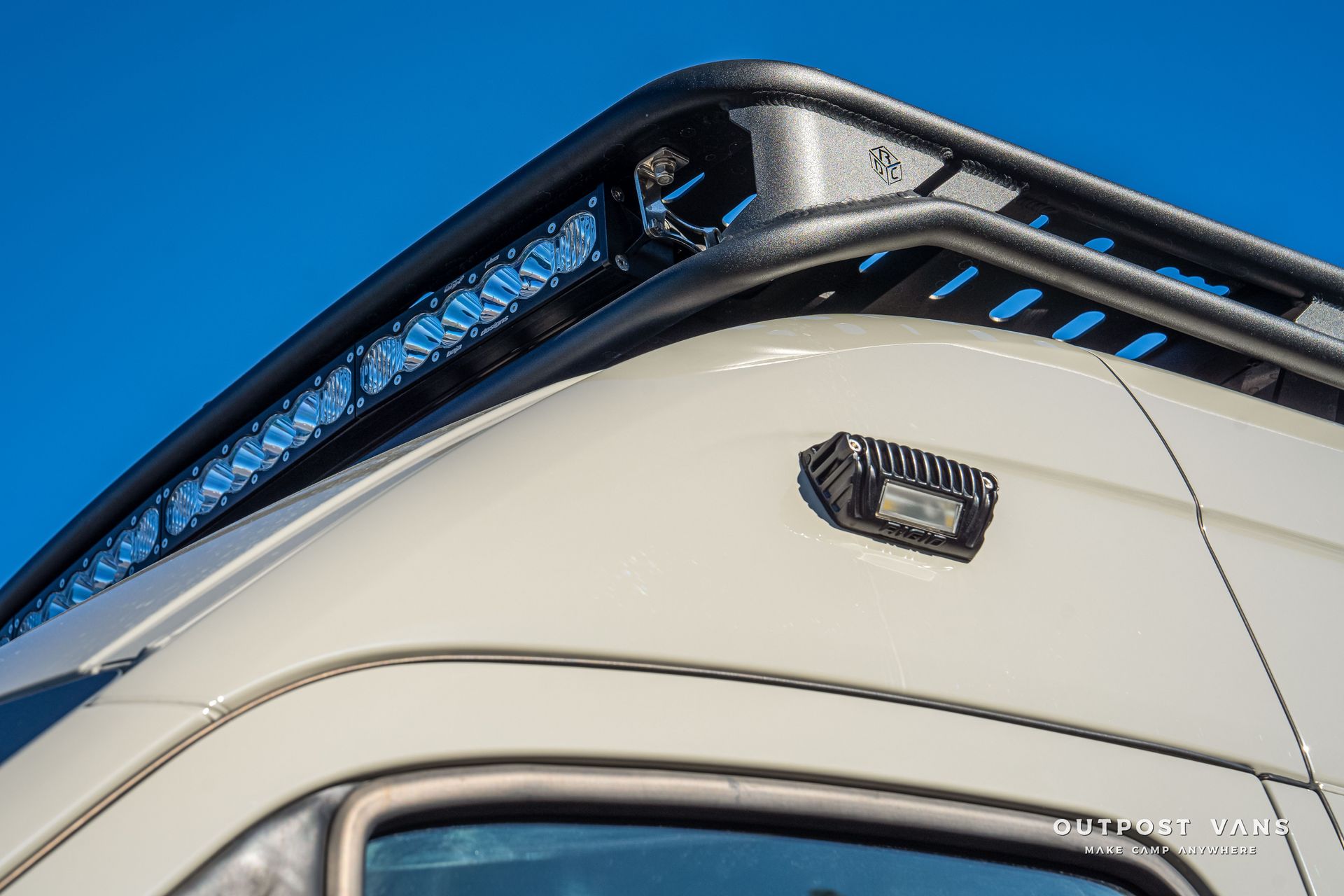 Off-white van corner with black roof rack and side-mounted light fixture against a clear blue sky.