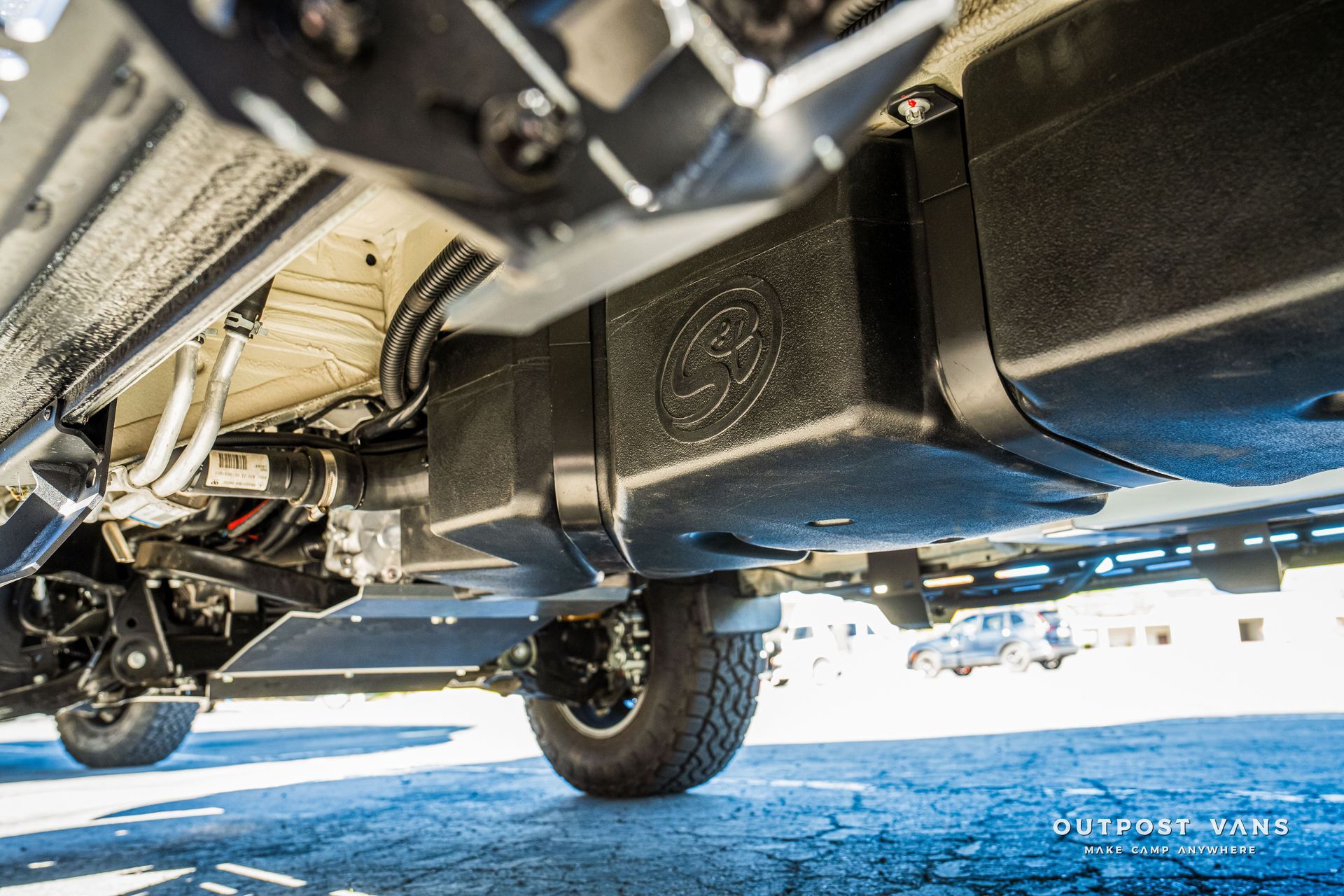Underside of a van, showing a black water tank and protective metal panels.