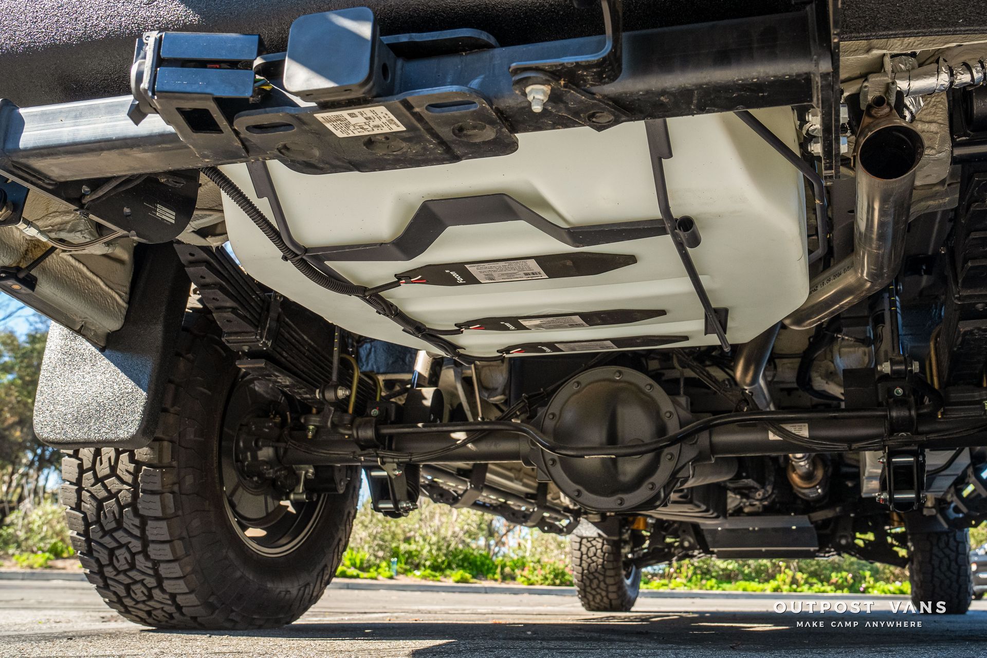 Underside of a vehicle showing rear axle, fuel tank, suspension, and tires on an outdoor surface.