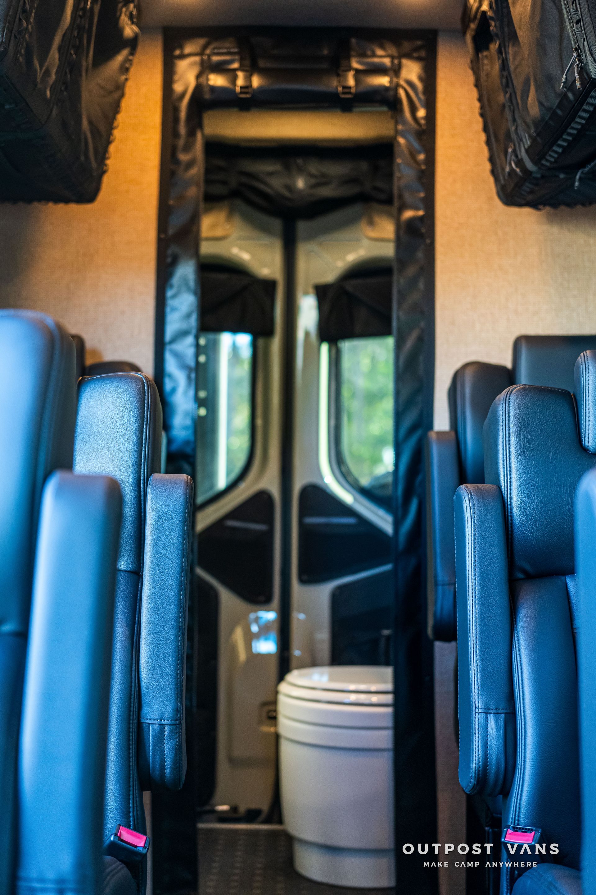 Interior view of a van, looking toward the open back doors. Seats line the sides, and a toilet is visible.