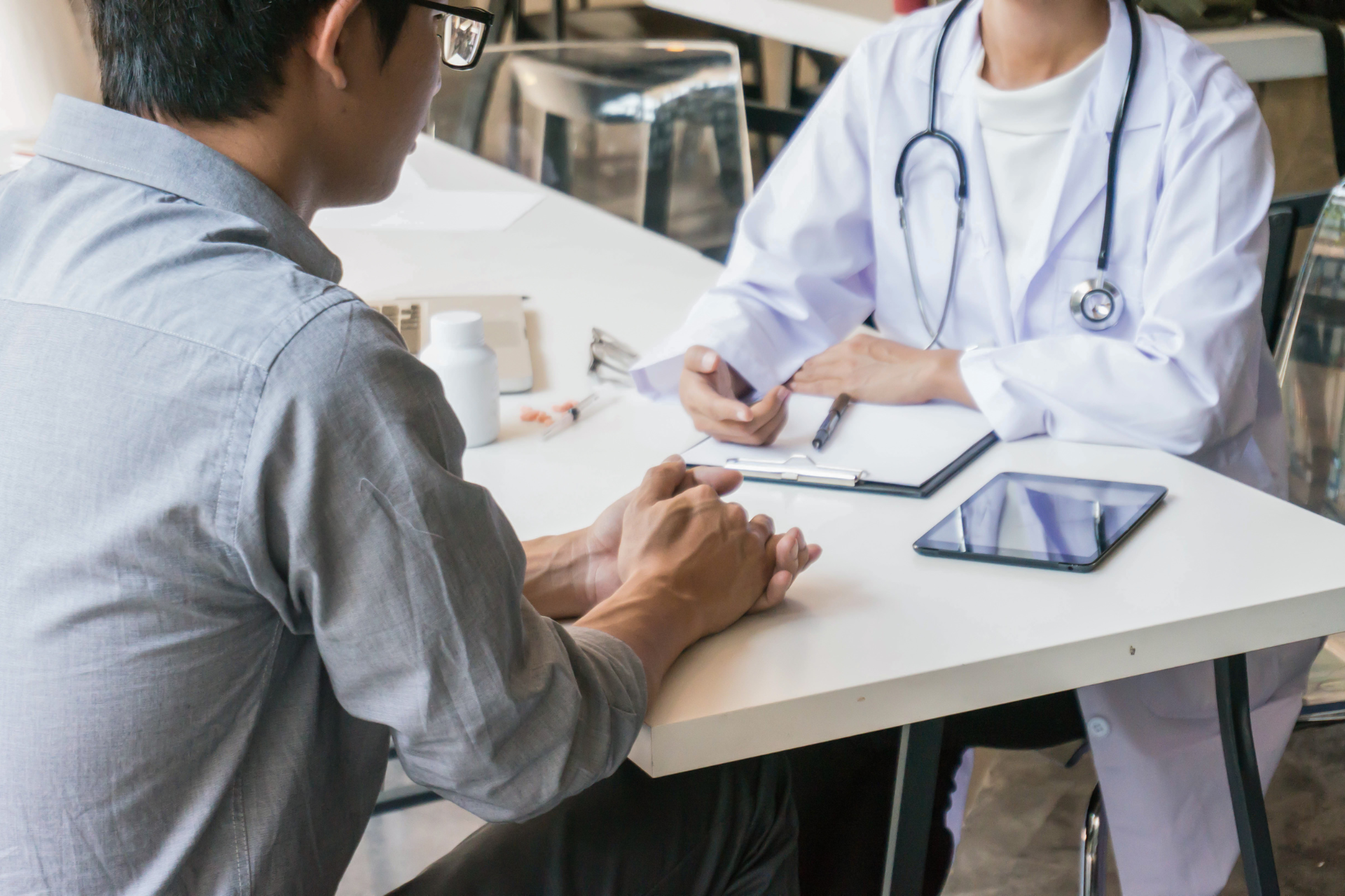 Patient consulting with a doctor at a table; tablet and paperwork visible in a clinic setting