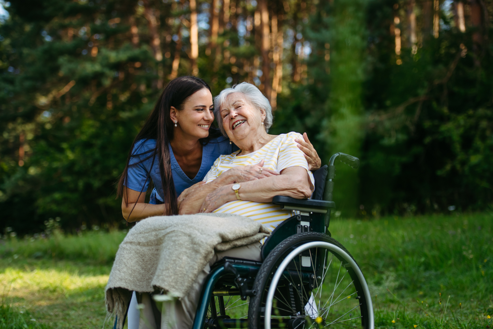 Woman in blue hugging an elderly person in a wheelchair in a sunny park
