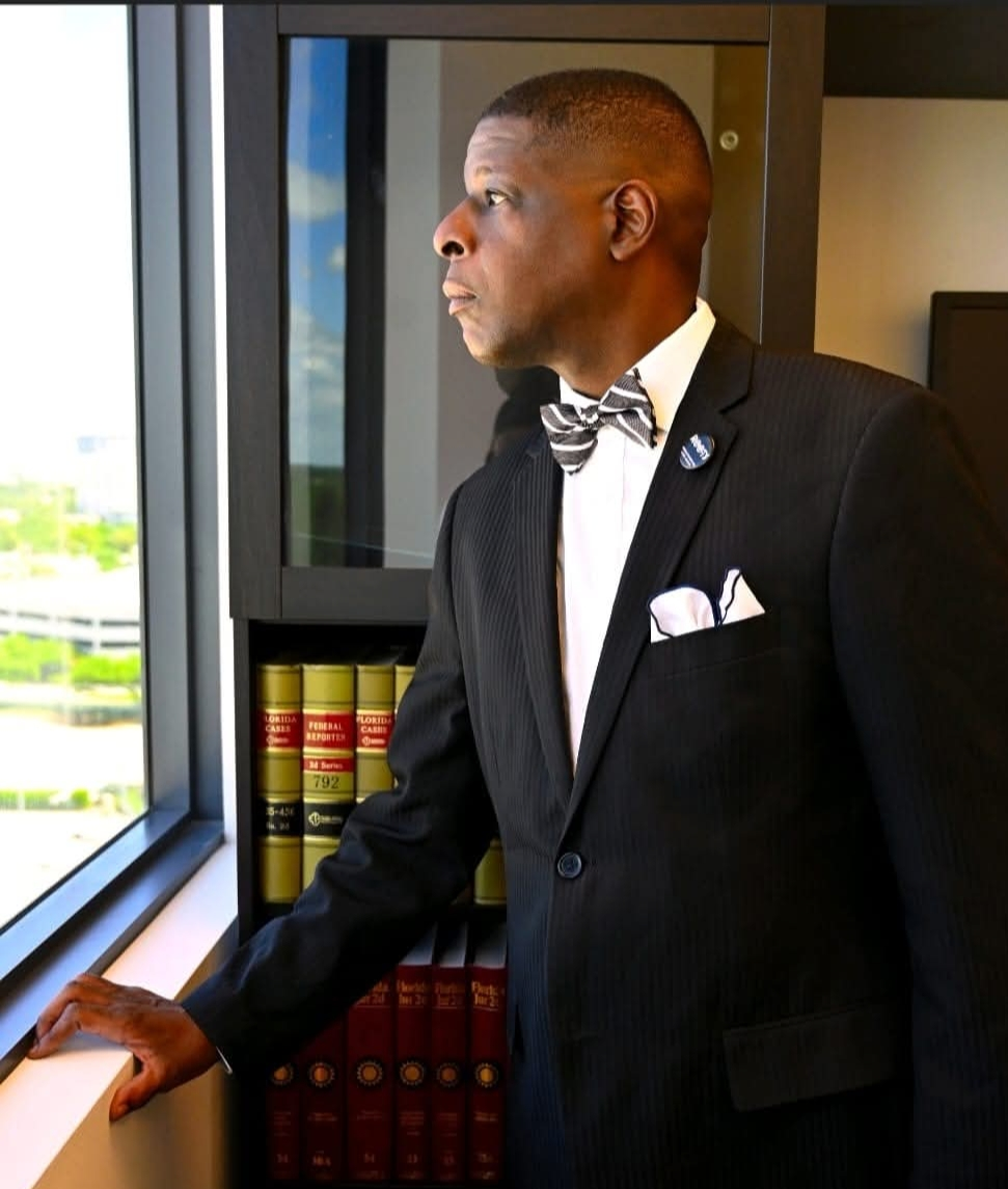 Law Office of Irvin Daphnis- Man in suit with bow tie, looking out window, arm resting. Bookshelf in background.