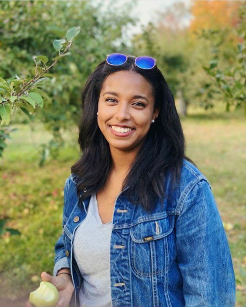 A smiling woman wearing a denim jacket and sunglasses is holding an apple.