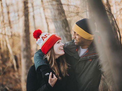 A man and a woman are standing next to each other in the woods and smiling at each other.