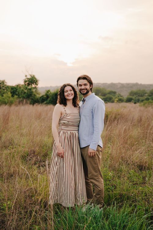 Couple standing together in a grassy field at sunset, smiling at the camera.