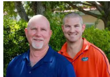 Two men smiling outdoors. One in blue, the other in orange. Green foliage in background.