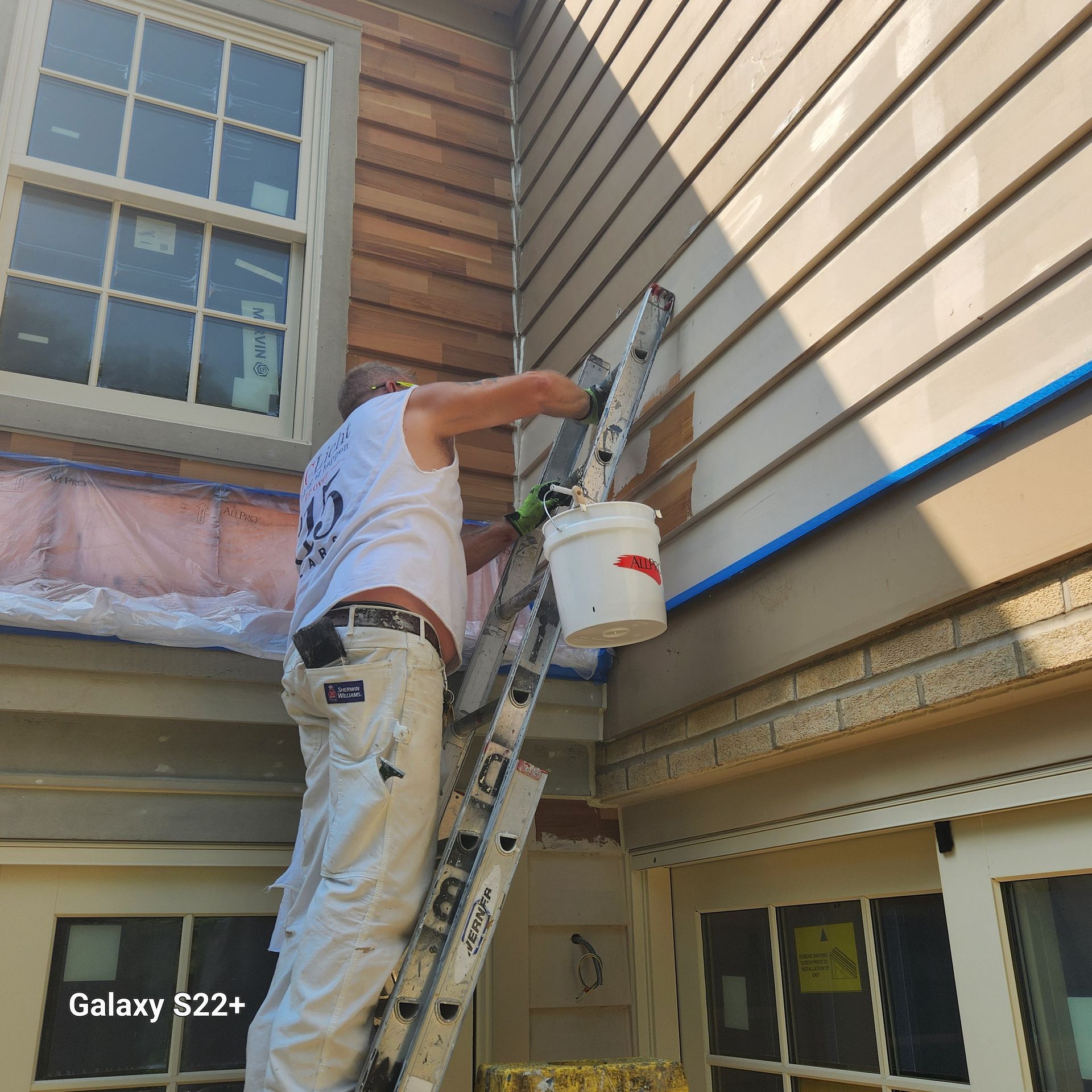 A man is standing on a ladder painting the side of a house.
