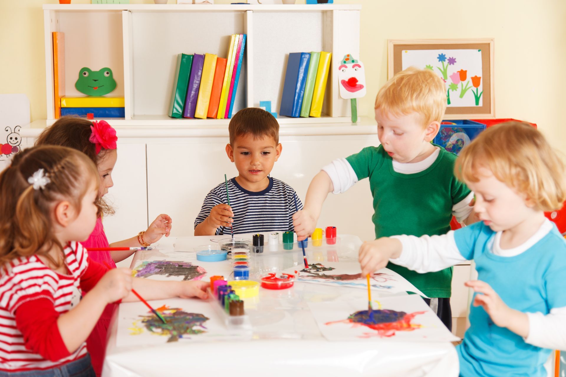Children painting together at a table in a bright classroom, with colorful art supplies and drawings.