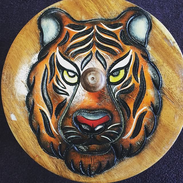 Tiger face carved and painted on a wooden circle; orange and black stripes, yellow eyes, red nose.