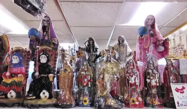 A collection of Santa Muerte statues in various colors and poses, displayed on a shelf indoors.