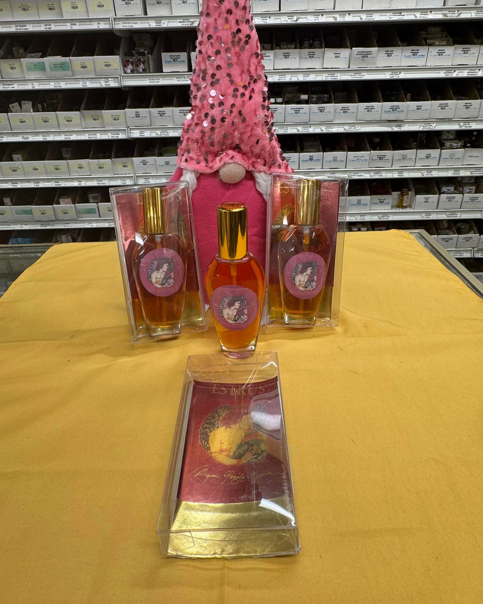 Bottles of perfume with a pink gnome on a yellow table. Chocolate bar in front. Background of shelves.