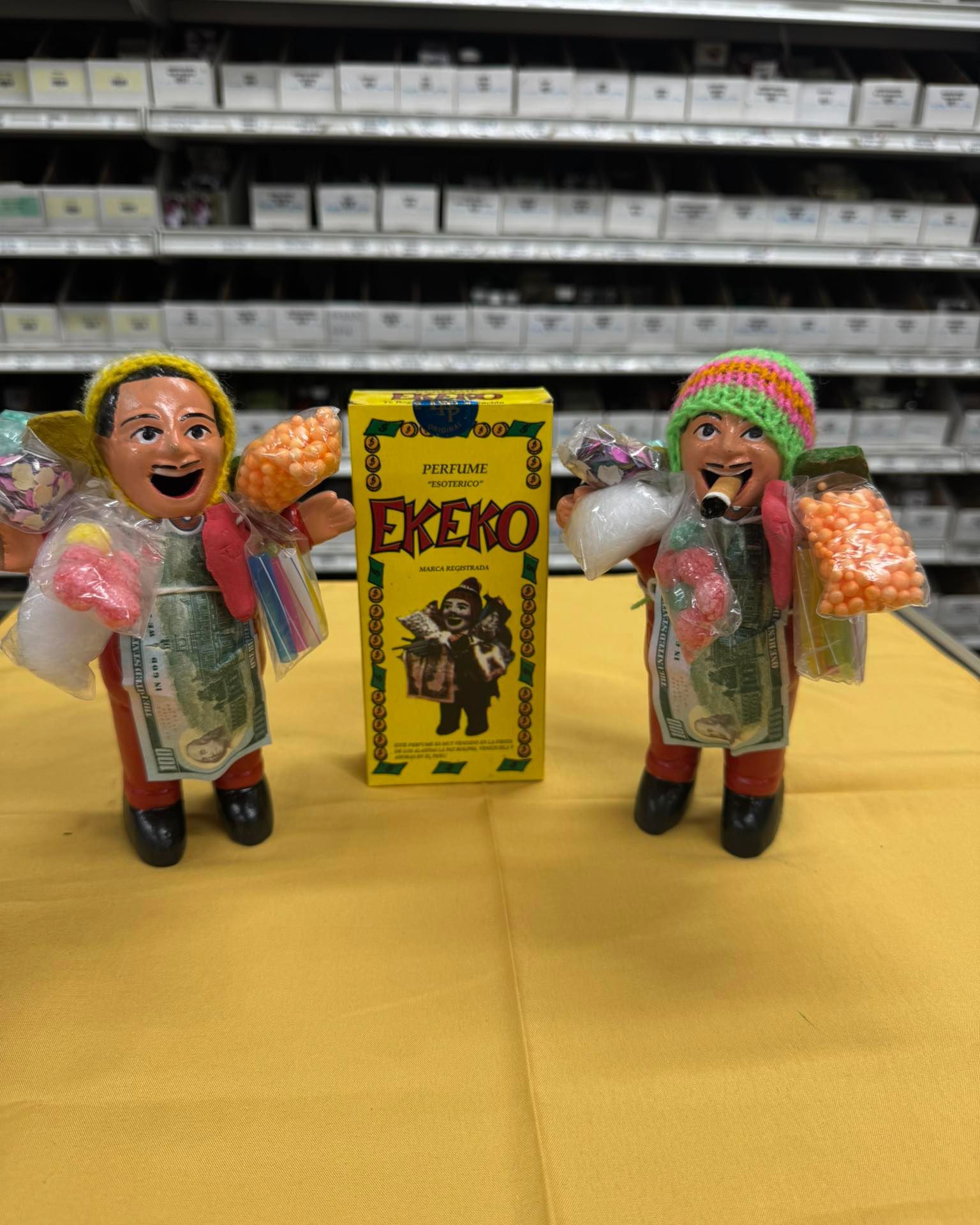 Two figurines flanking a yellow box. Figurines have smiling faces, holding items and wearing colorful hats. Shelves are in background.