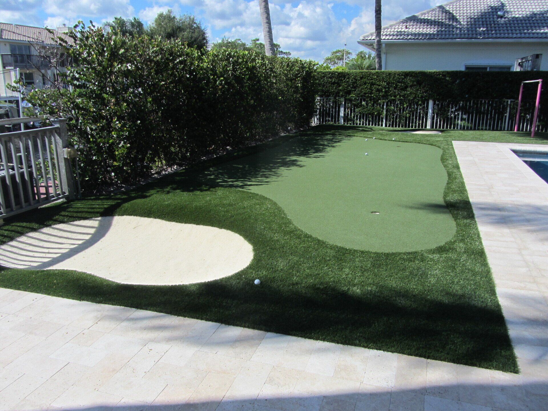 Backyard putting green with sand trap, surrounded by hedges, next to a pool.