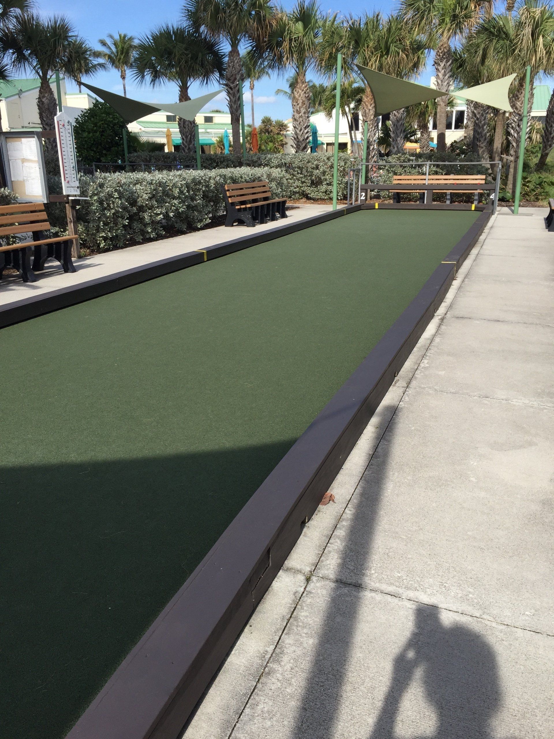 Bocce ball court with green turf, benches, and shade structures; outdoors on a sunny day.