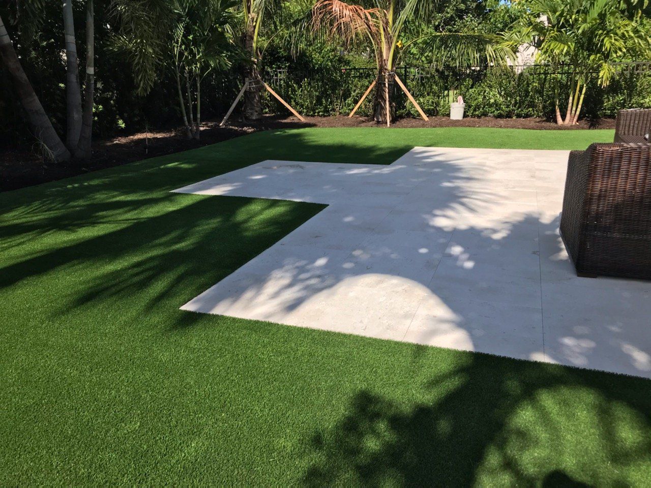 Green artificial grass surrounds a light concrete patio in a backyard setting with trees.