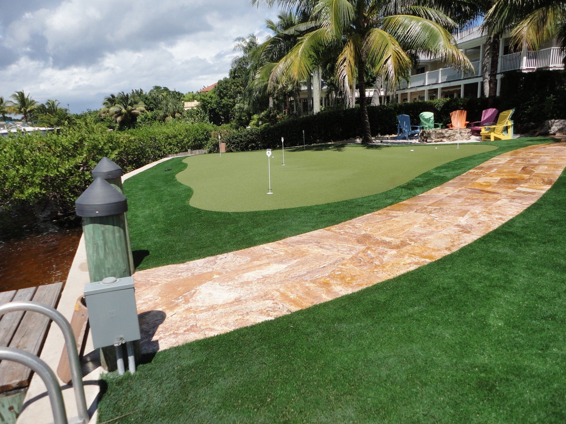 A putting green with a stone path. Palm trees and colorful chairs are in the background near water.