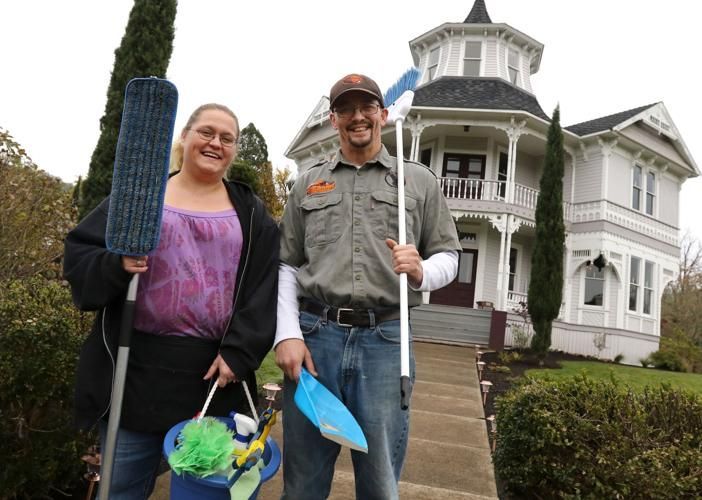 Two people with cleaning supplies stand in front of a Victorian house; woman holds a mop, man a broom.
