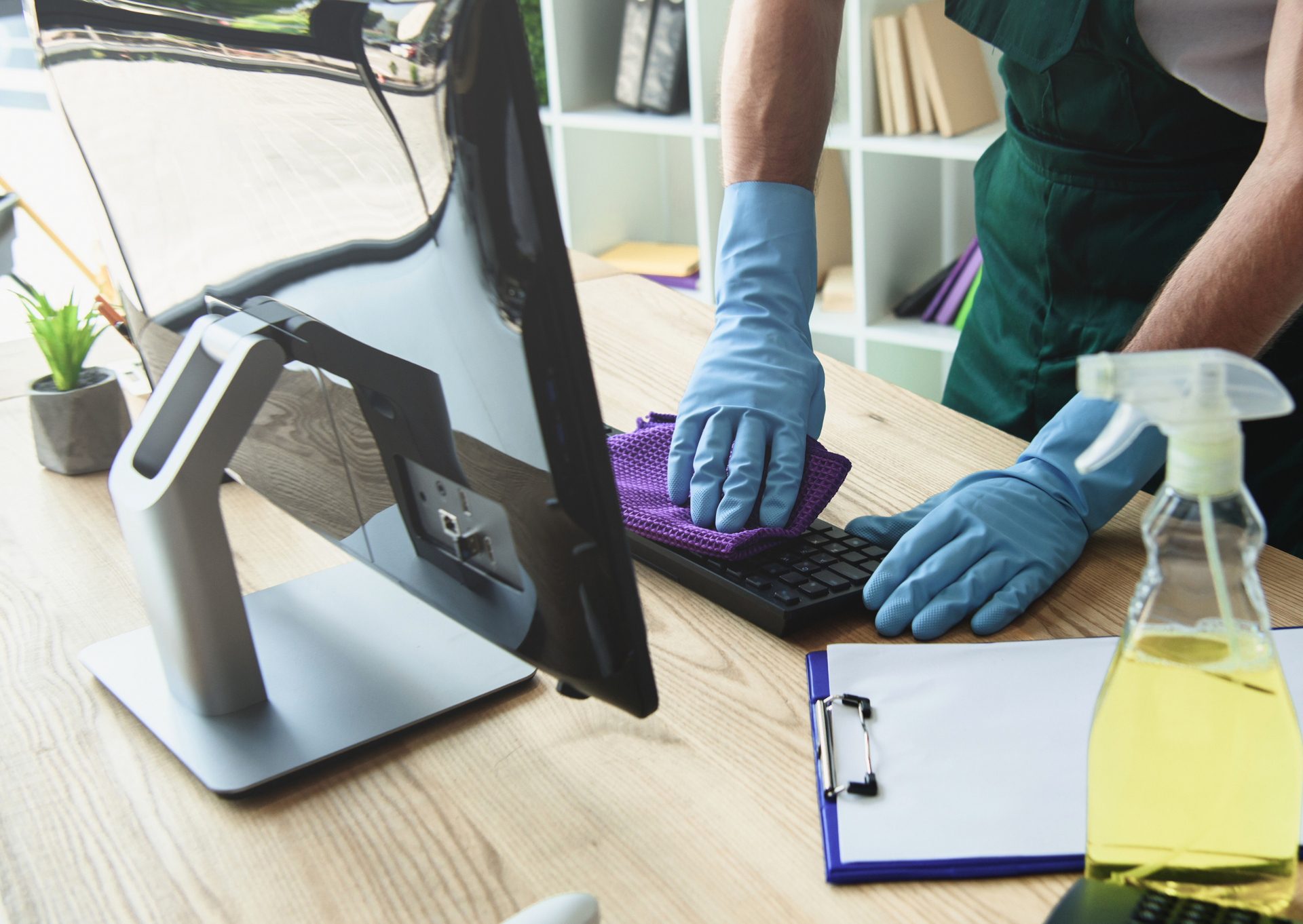 Person wearing gloves cleaning a computer keyboard with a spray bottle on a desk in an office.