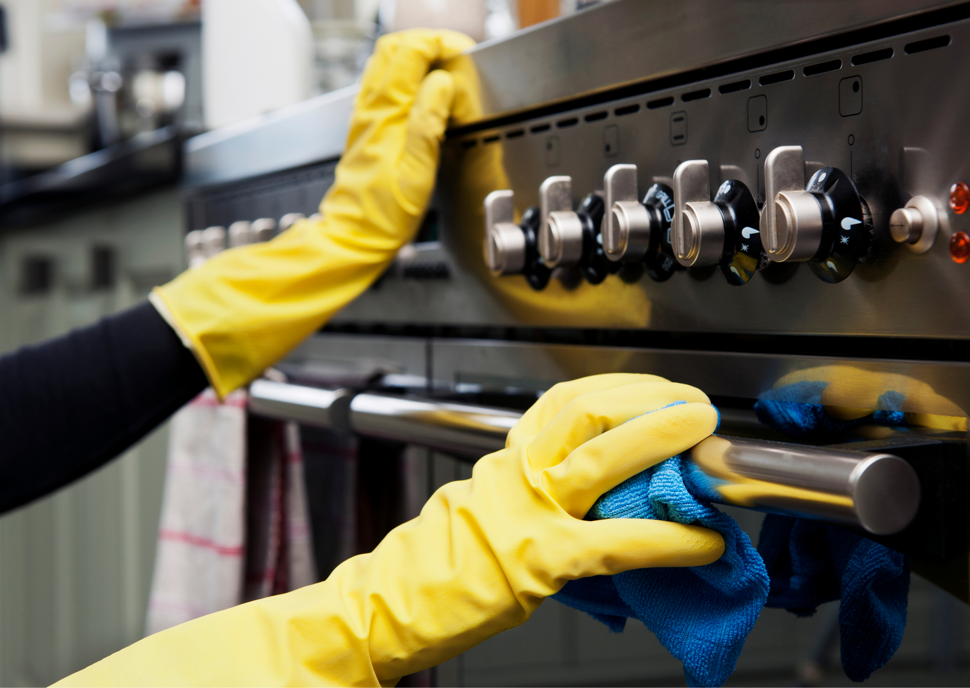 Hands in yellow gloves cleaning an oven with a blue cloth. Kitchen setting.