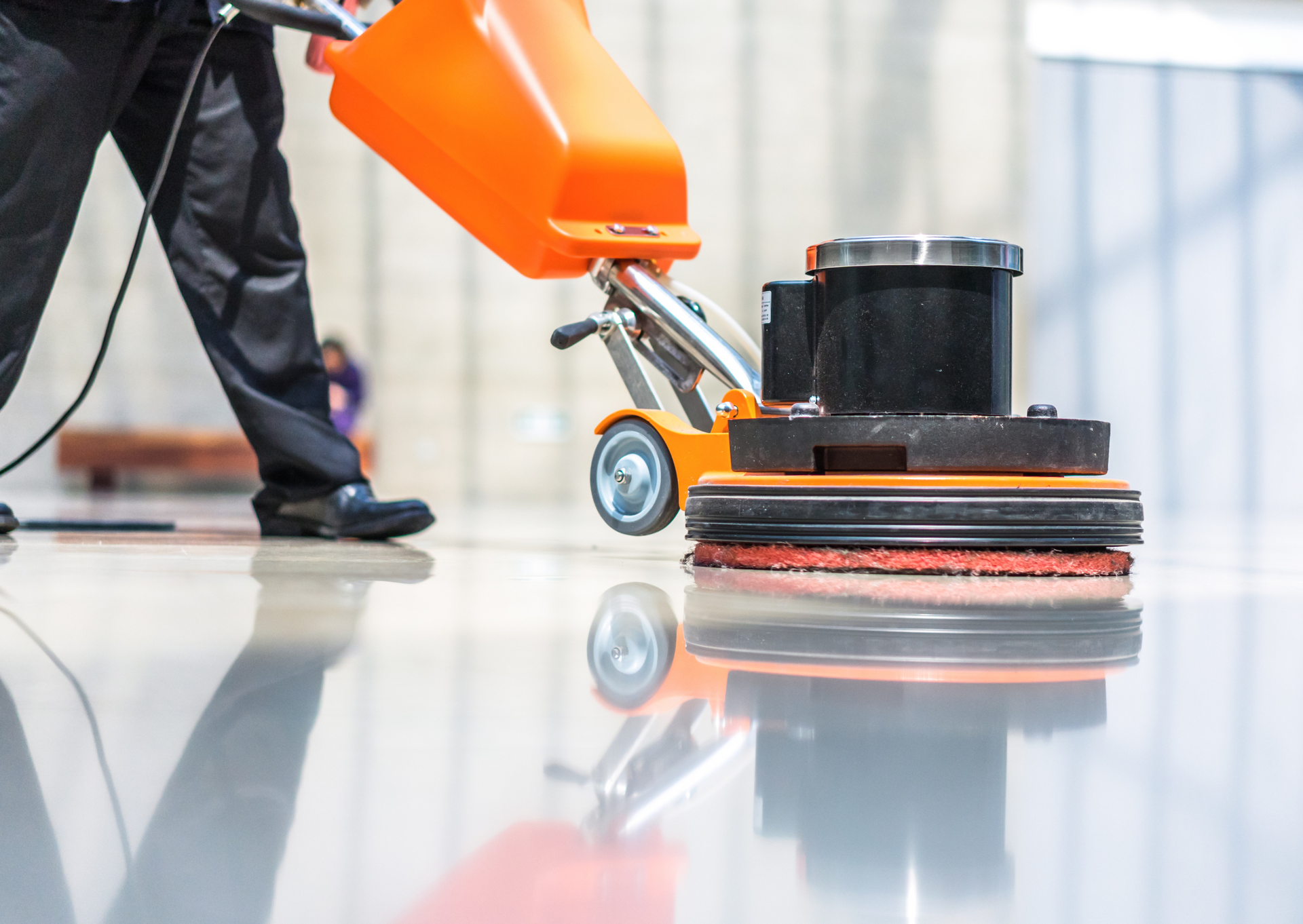 Person cleaning a shiny floor with an orange and black floor buffer machine.