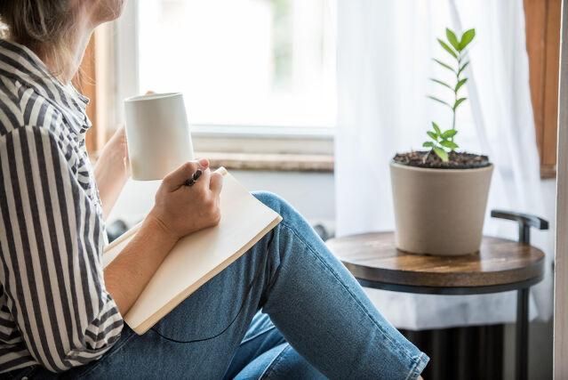 Woman sitting writing on a notepad in front of a window next to a potted plant