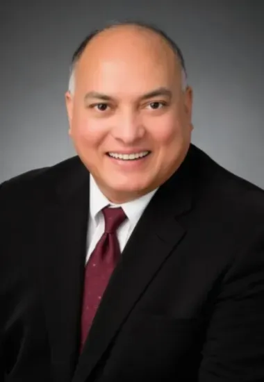 Man in a suit smiles, dark tie, against a gray backdrop.