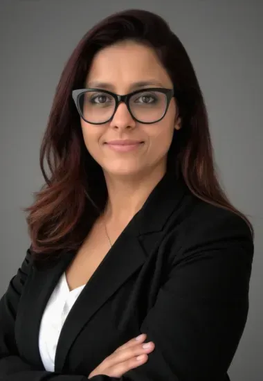 Woman in a black blazer and glasses, arms crossed, smiling, against a gray background.
