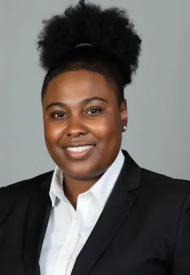 Woman with dark skin wearing a white shirt and black suit smiles at the camera, with natural hair up.