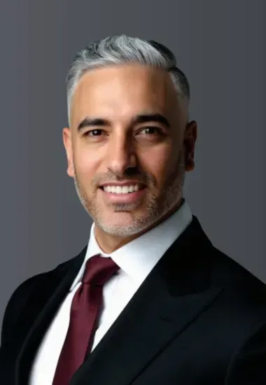 Man in dark suit smiles, wearing a burgundy tie. Gray hair, studio setting.