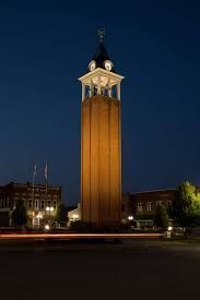 Tall brick clock tower at night, illuminated, with buildings and dark sky in background. Marion, Il. Marion town square. downtown marion, il