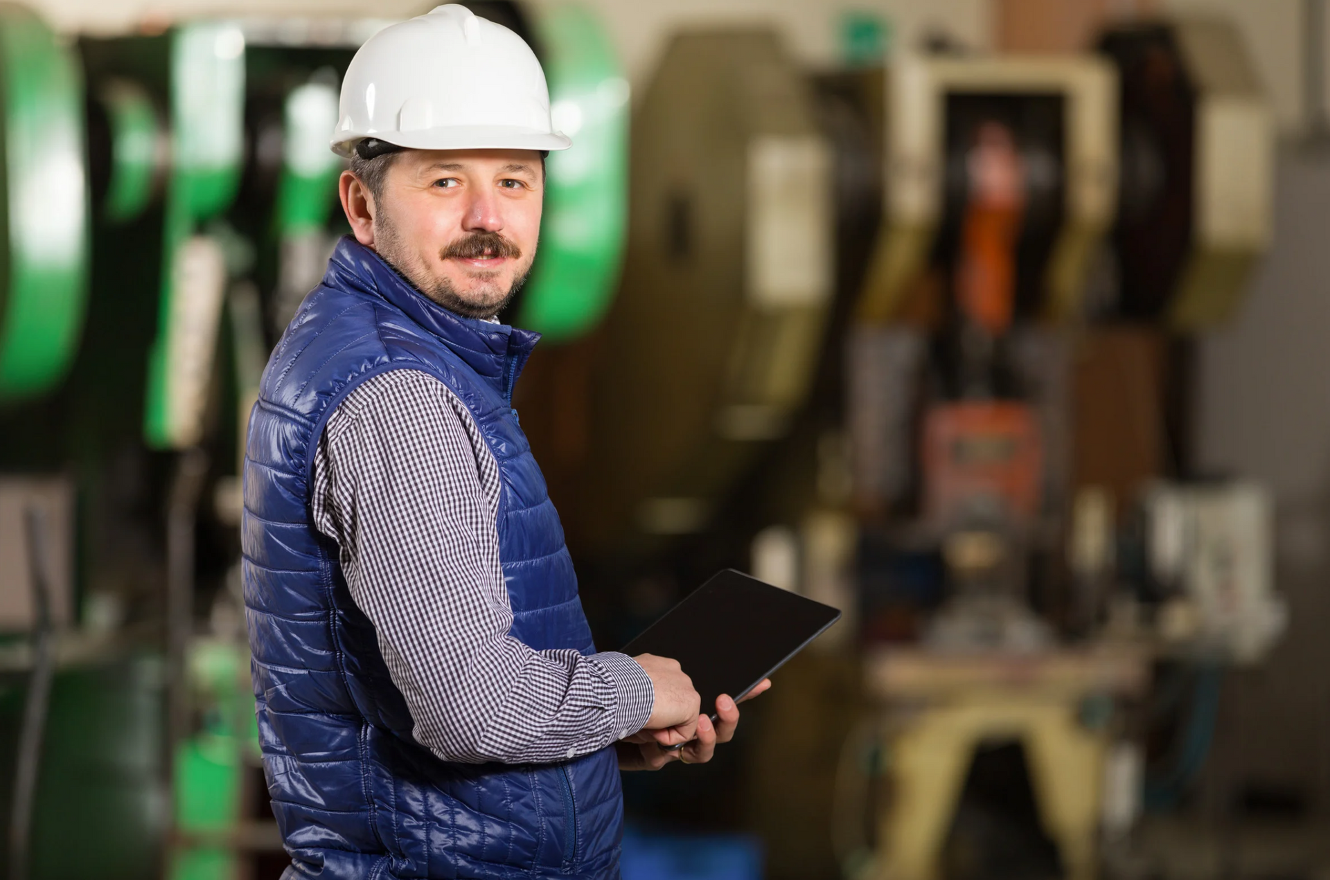 Man in white hard hat and vest holding tablet, in a factory setting.